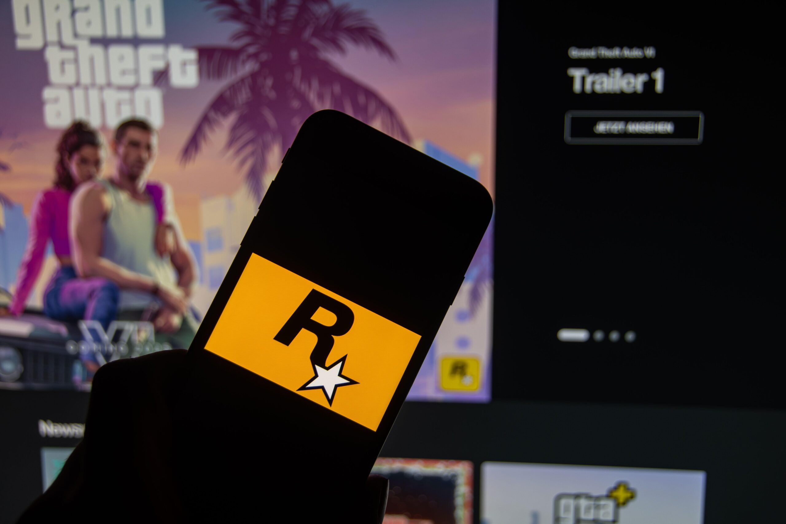 a person holding a phone with the Rockstar logo with GTA VI trailer in the background on a monitor
